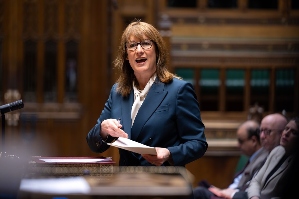 A woman in glasses and a dark blue suit speaking at a podium with papers in hand.