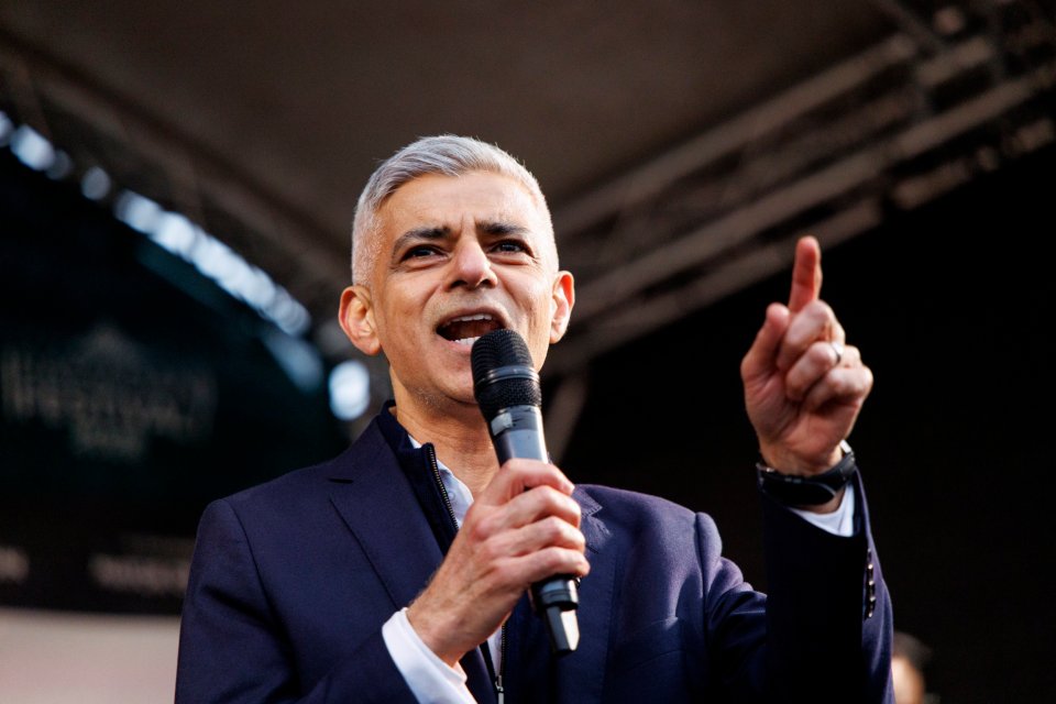 Sadiq Khan speaking into a microphone, with his right hand raised and index finger pointing up.