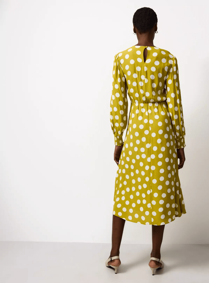 Rear view of a model wearing a knee-length olive green dress with white polka dots and kitten heels.