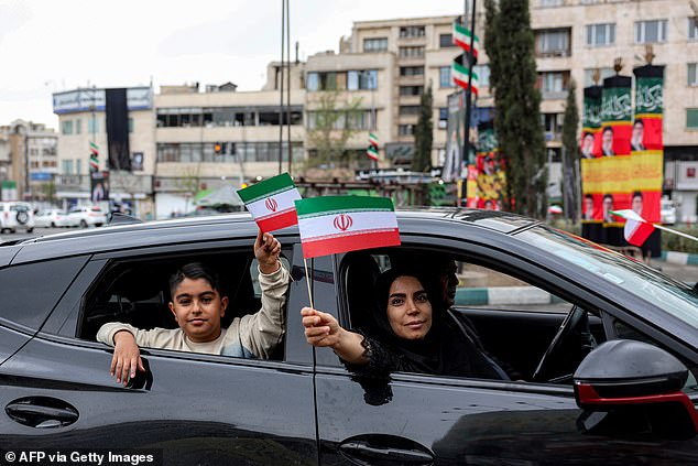 A woman and boy hold up miniature Iranian flags while sitting in a vehicle moving along Enghelab (Revolution) Square in central Tehran on March 25, 2026