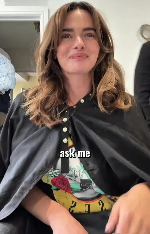 A woman with long brown hair, wearing a black cape over a Guns N' Roses t-shirt, smiles at the camera.