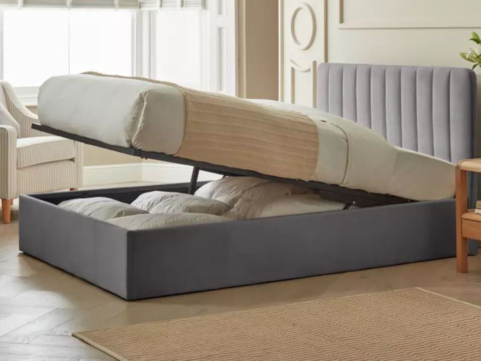A gray storage bed with the mattress lifted to reveal pillows and blankets stored inside.