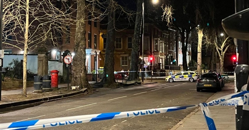 Police tape blocking off a city street at night, with a police car and other vehicles visible in the background.