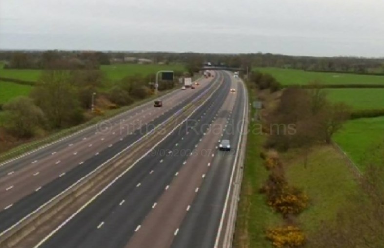 Overhead view of a highway with moderate traffic, surrounded by green fields under an overcast sky.