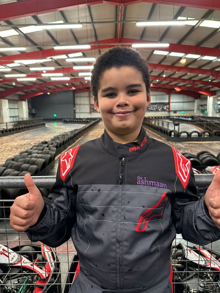 Theo Bobb, a young karter, gives two thumbs up while wearing a racing suit on an indoor karting track.