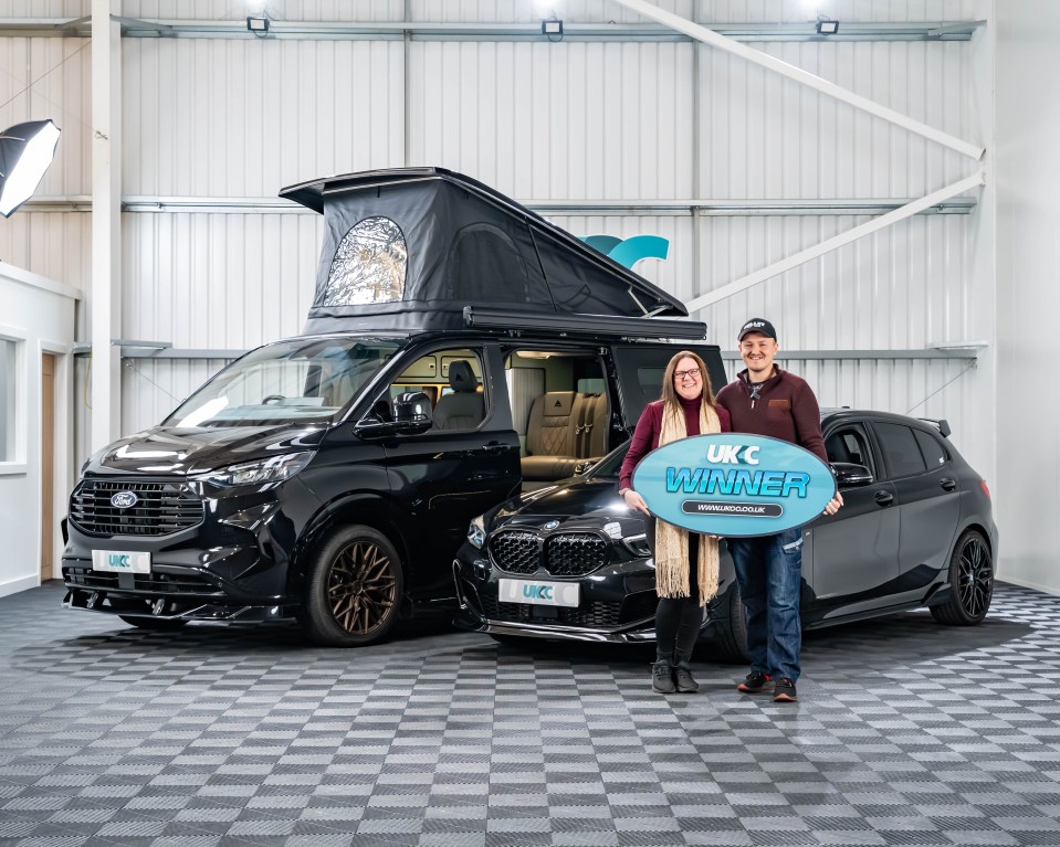 A man and a woman standing in front of a black camper van and a black car, holding a blue sign that says "UKC WINNER".