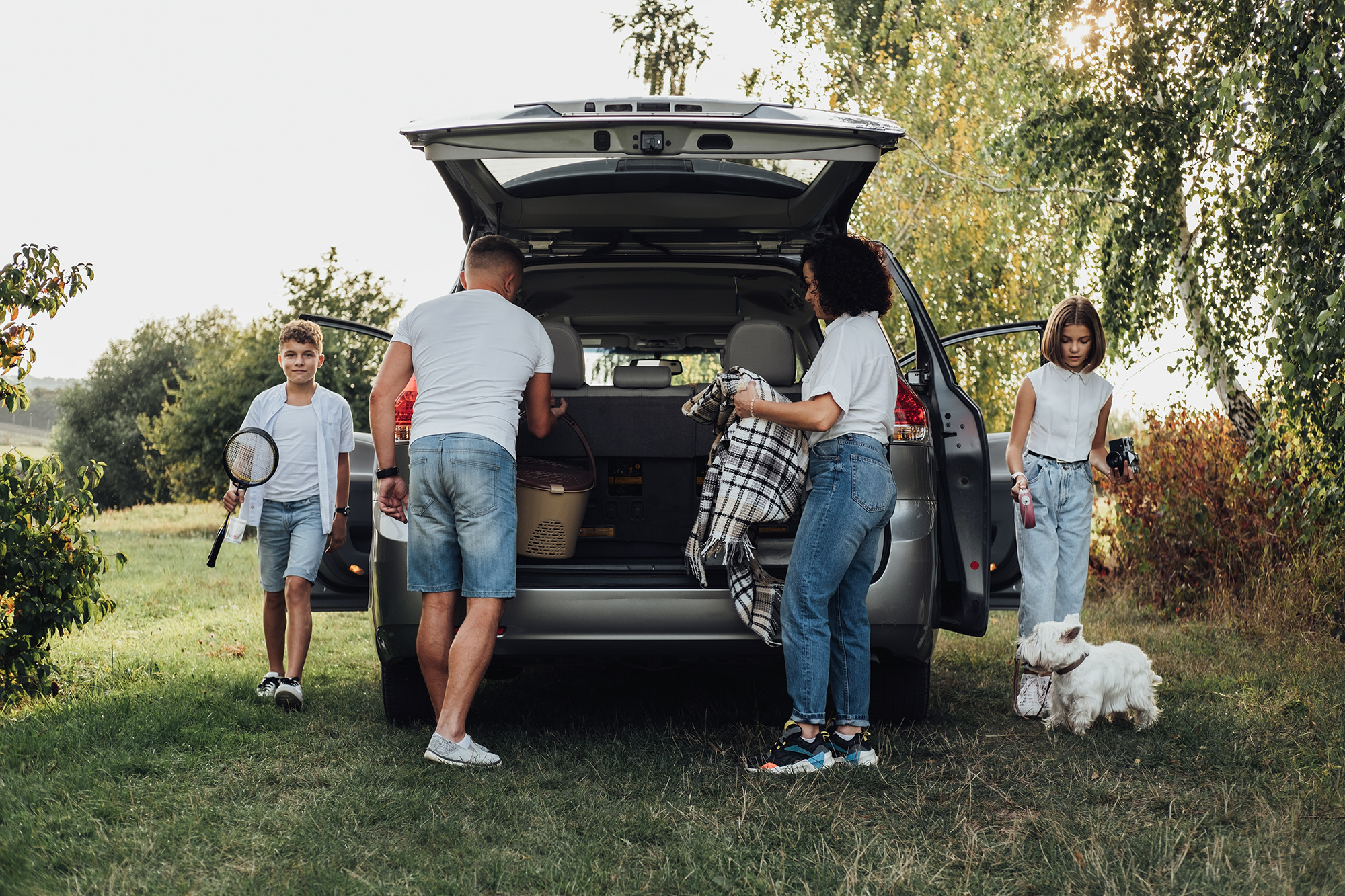 A family of four and their dog unpacking a car for a picnic.