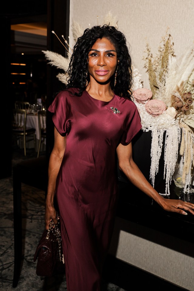 Sinitta in a maroon satin gown with a brooch, holding a matching quilted leather purse.