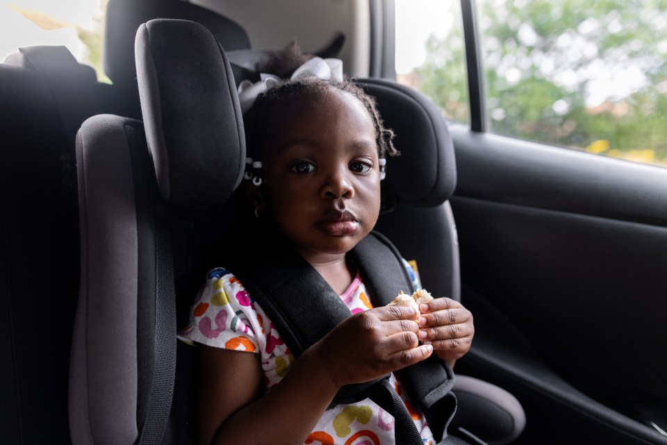 A young girl in a car seat eating a sandwich in the back of an electric vehicle.