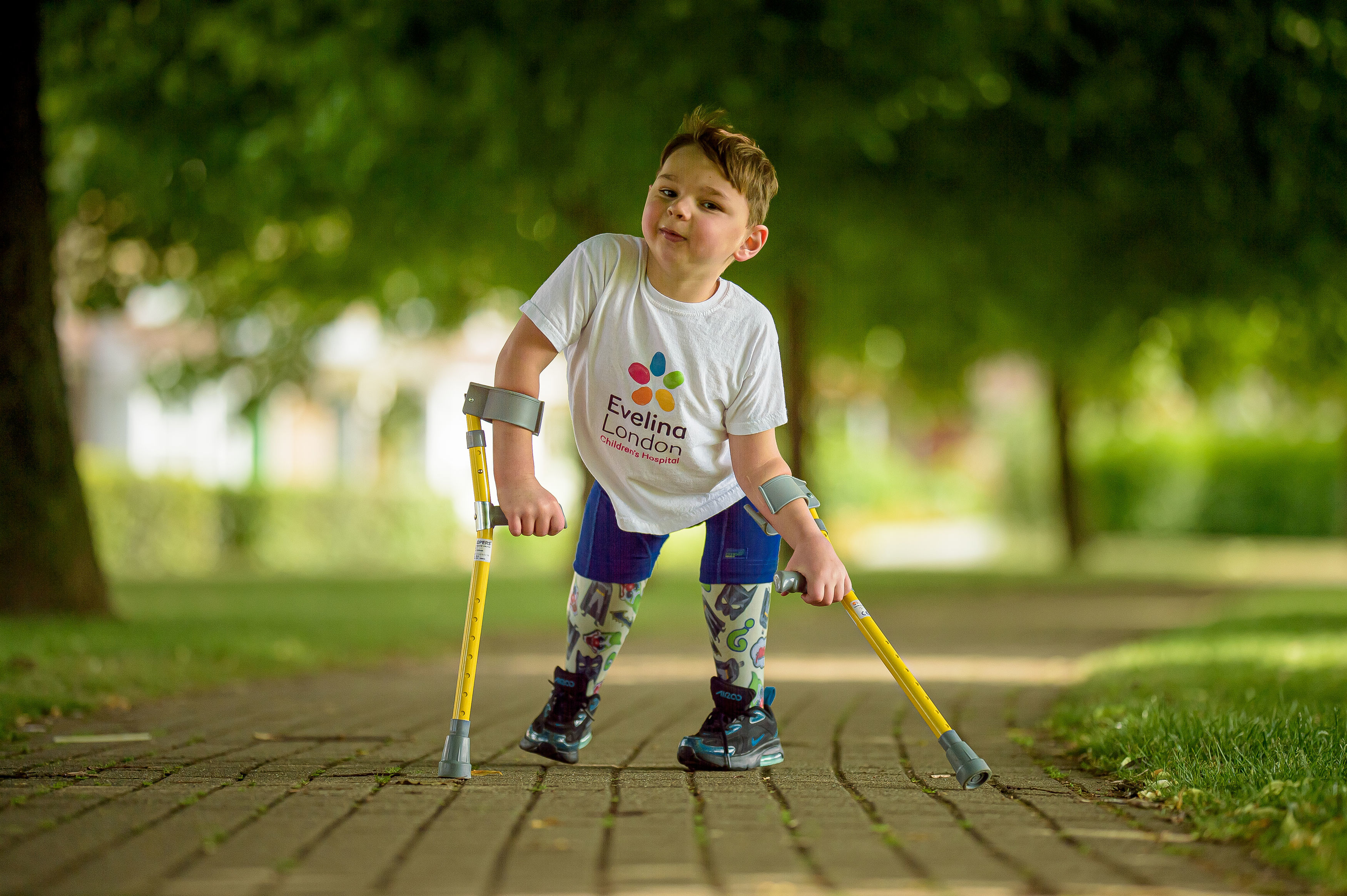 A young boy with prosthetic legs and arm crutches wears a white t-shirt with the logo "Evelina London Children's Hospital" and smiles at the camera while walking along a paved path with green grass and trees in the background.
