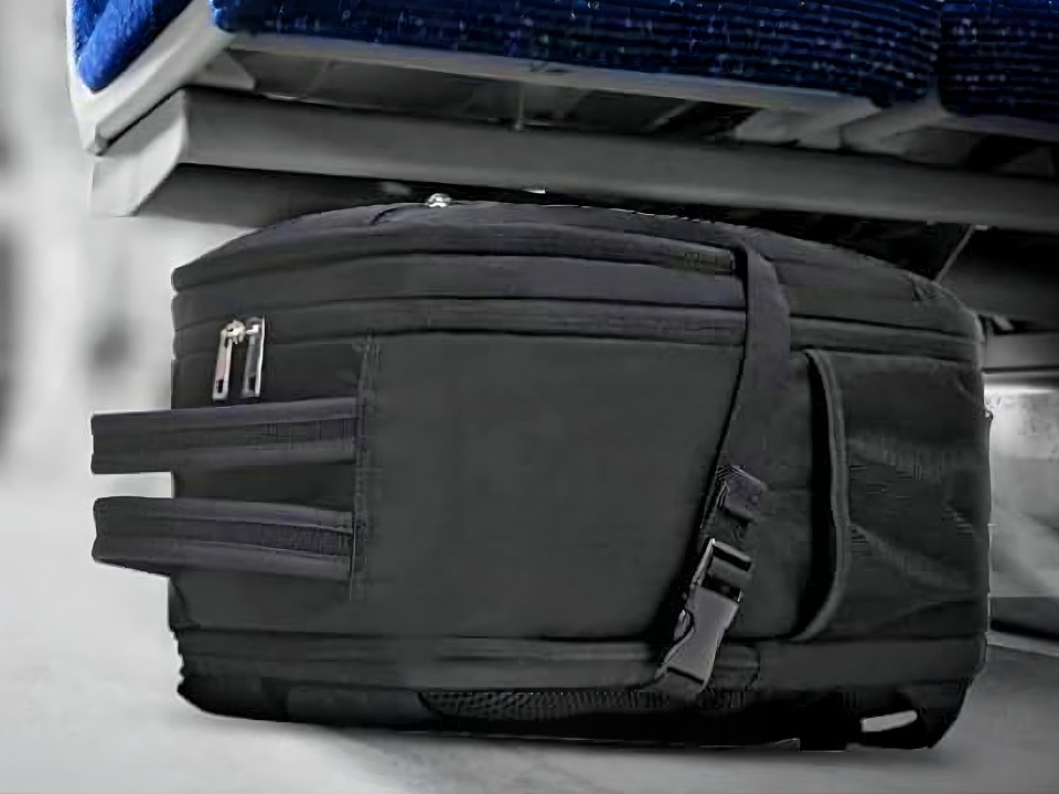 A black underseat cabin bag with a side pocket and compression straps, tucked under a blue airline seat.