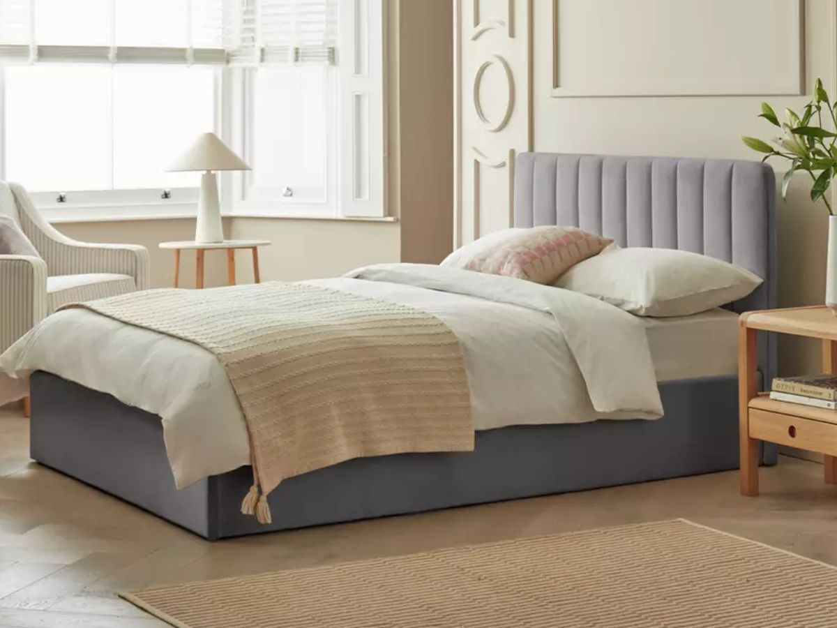 A bed with a gray upholstered headboard and base, white duvet, and beige knitted throw in a bedroom with a large window and wooden nightstand.