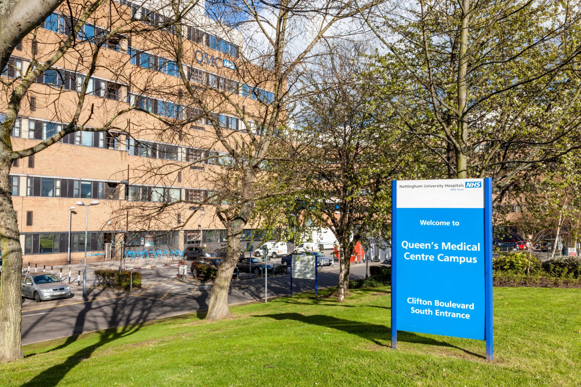 An image collage containing 1 images, Image 1 shows The Queen's Medical Centre Nottingham, a large brick hospital building, with a blue sign in the foreground welcoming visitors to the campus via the Clifton Boulevard South Entrance