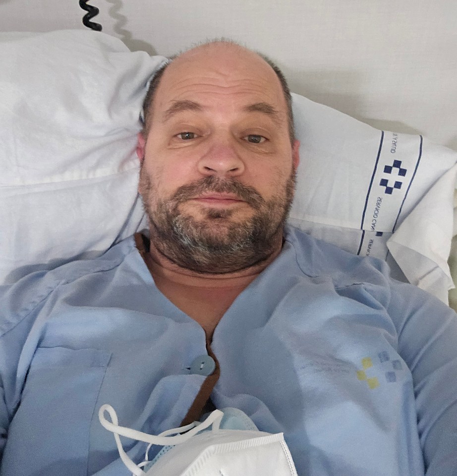 A man in a hospital gown lies in bed, looking directly at the camera.
