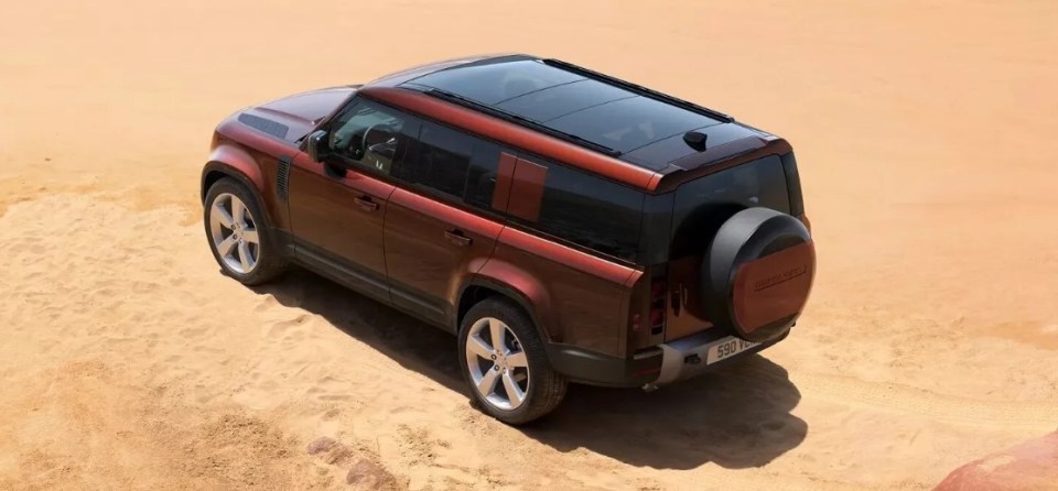Brown Land Rover Defender with a black panoramic roof, parked in sand.