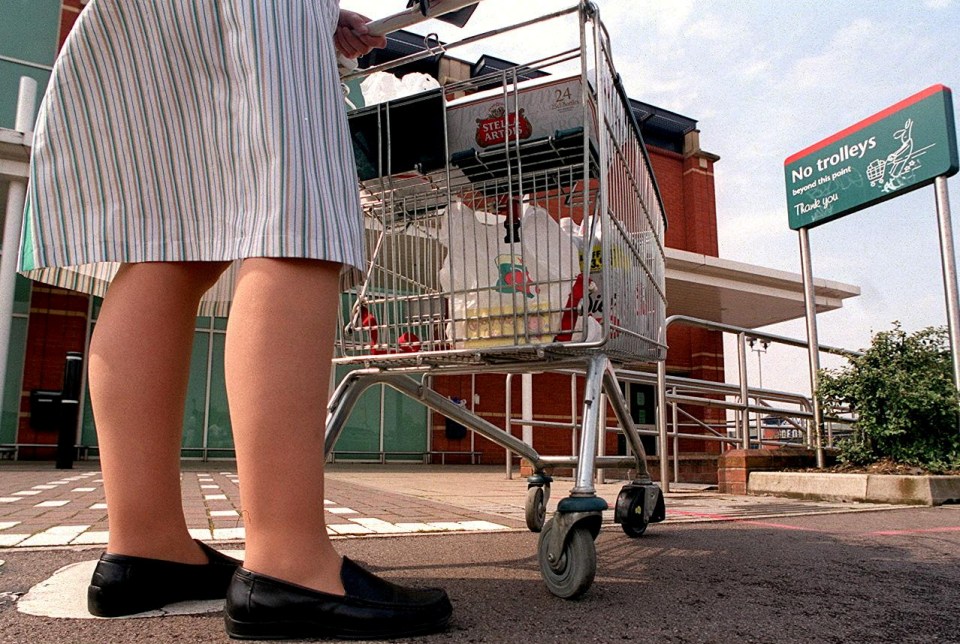 A person pushing a shopping cart full of groceries, with a sign in the background reading "No trolleys beyond this point."