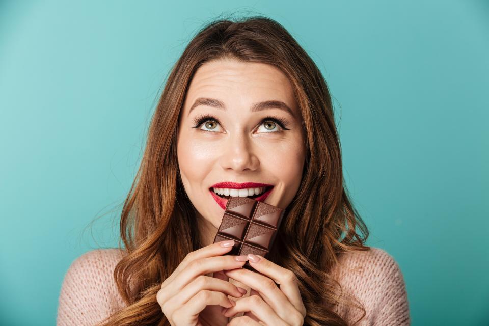 Portrait of a delighted brown haired woman with bright makeup eating chocolate bar isolated over blue background