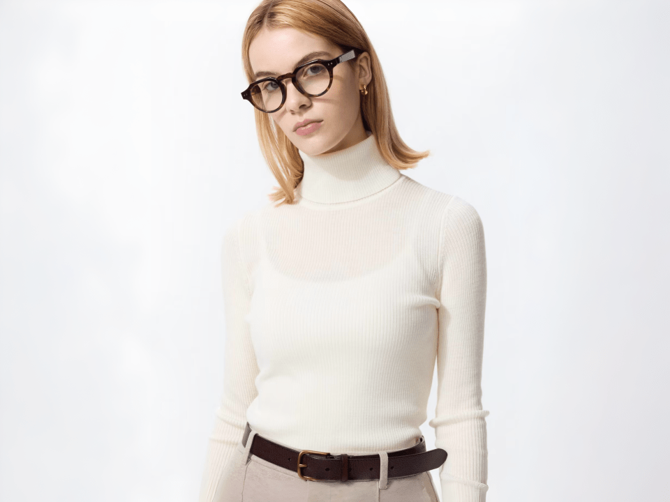 A woman wearing a white ribbed turtleneck sweater, light-colored pants, a brown belt, and round glasses.