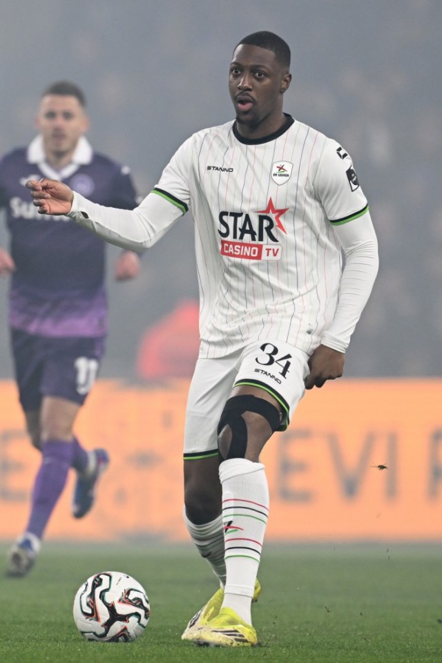 A football player in a white jersey and shorts with black and red stripes, number 34, stands on a green field next to a soccer ball.