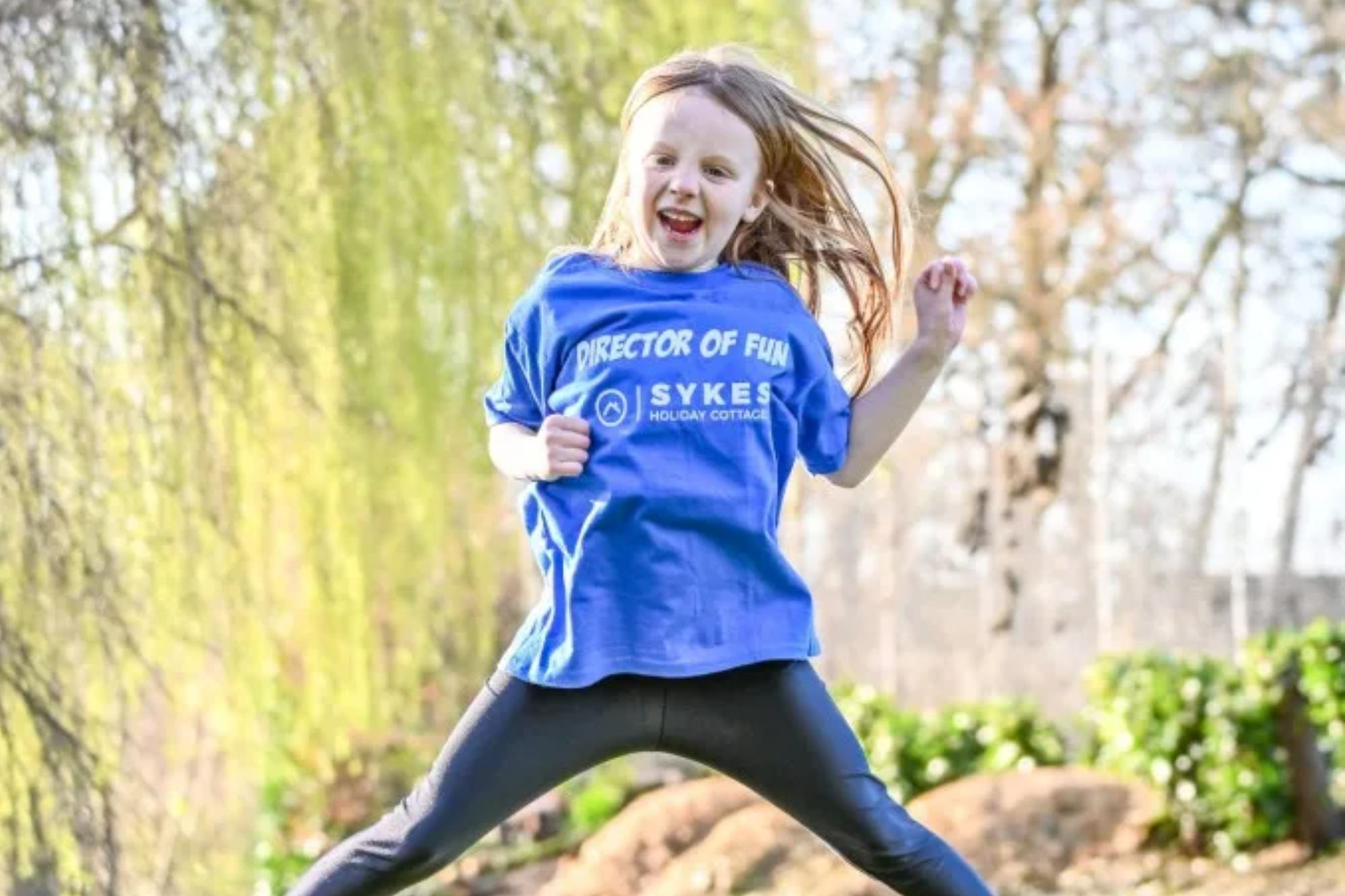 An image collage containing 1 images, Image 1 shows A young girl in a blue "Director of Fun" t-shirt jumping in the air in a park