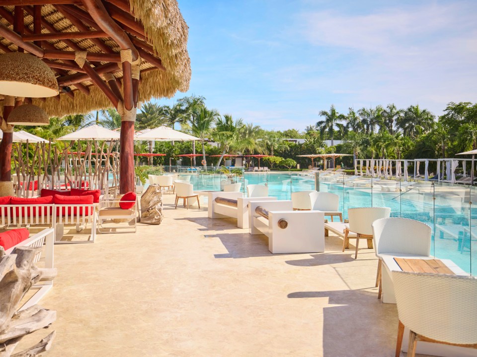 An outdoor seating area with red cushions under a thatched roof, next to a large swimming pool lined with palm trees.
