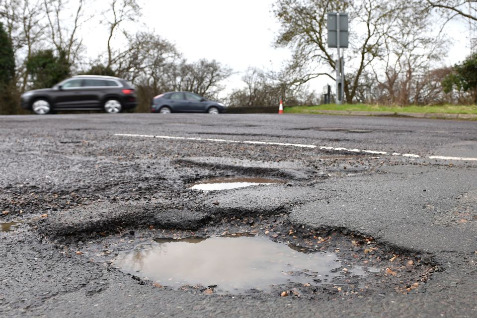 A close-up of several potholes filled with water on an asphalt road with cars blurred in the background.