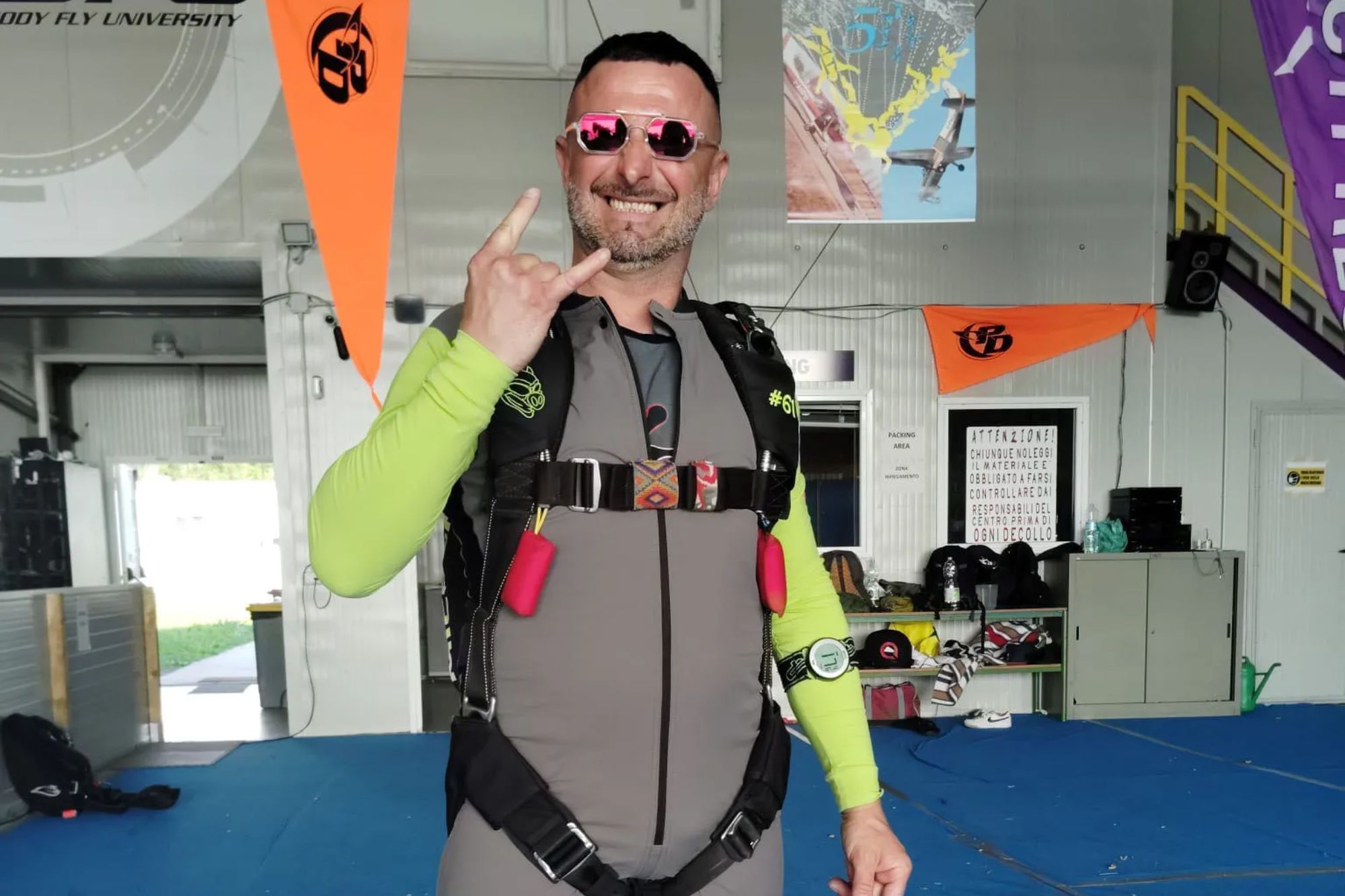 An image collage containing 1 images, Image 1 shows Simone Bacchiega in his skydiver suit, smiling and making a "rock on" hand gesture