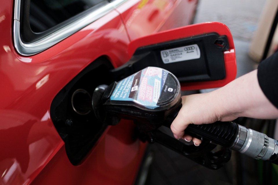 A person's hand holding a diesel pump nozzle in the fuel tank of a red car.