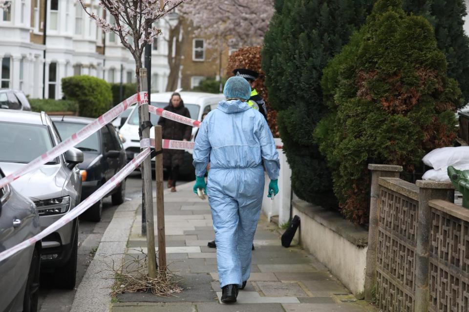 A forensic officer in a blue suit and green gloves walks down a sidewalk past a police barrier.