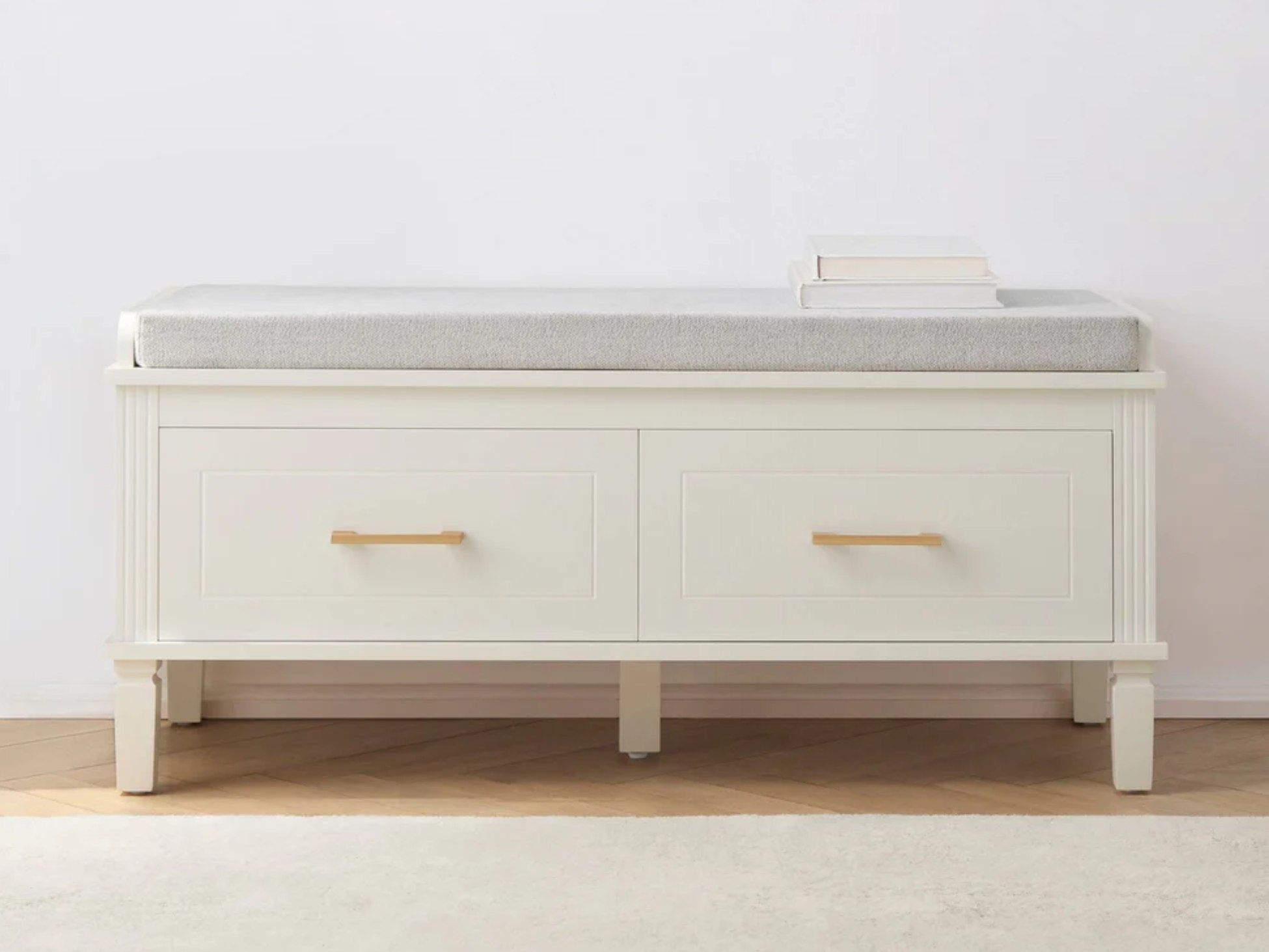 White storage bench with two drawers and a gray upholstered cushion on top, with a stack of white books on the right side of the cushion.