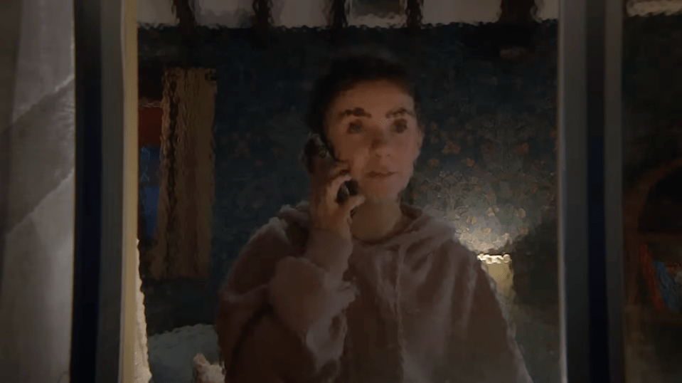 A young woman in a pink hoodie talking on a phone, seen through a textured window.