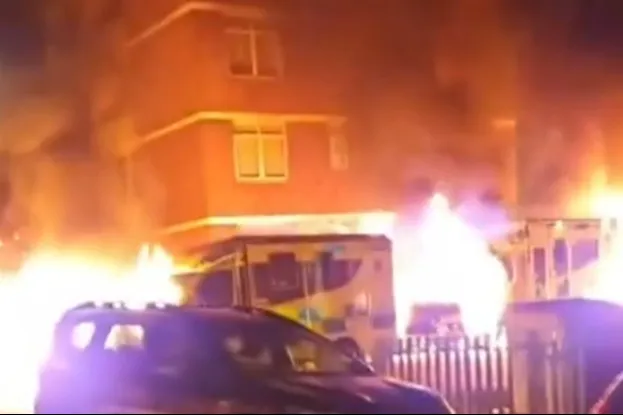 A building and an ambulance engulfed in flames, with cars in the foreground.