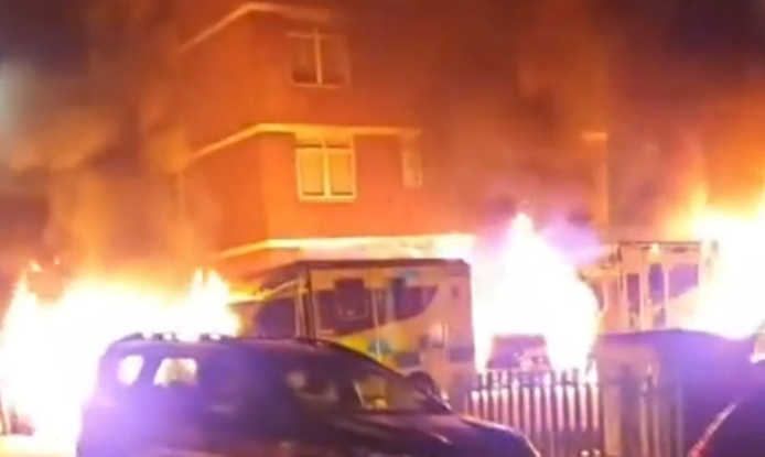 A building and an ambulance engulfed in flames, with cars in the foreground.