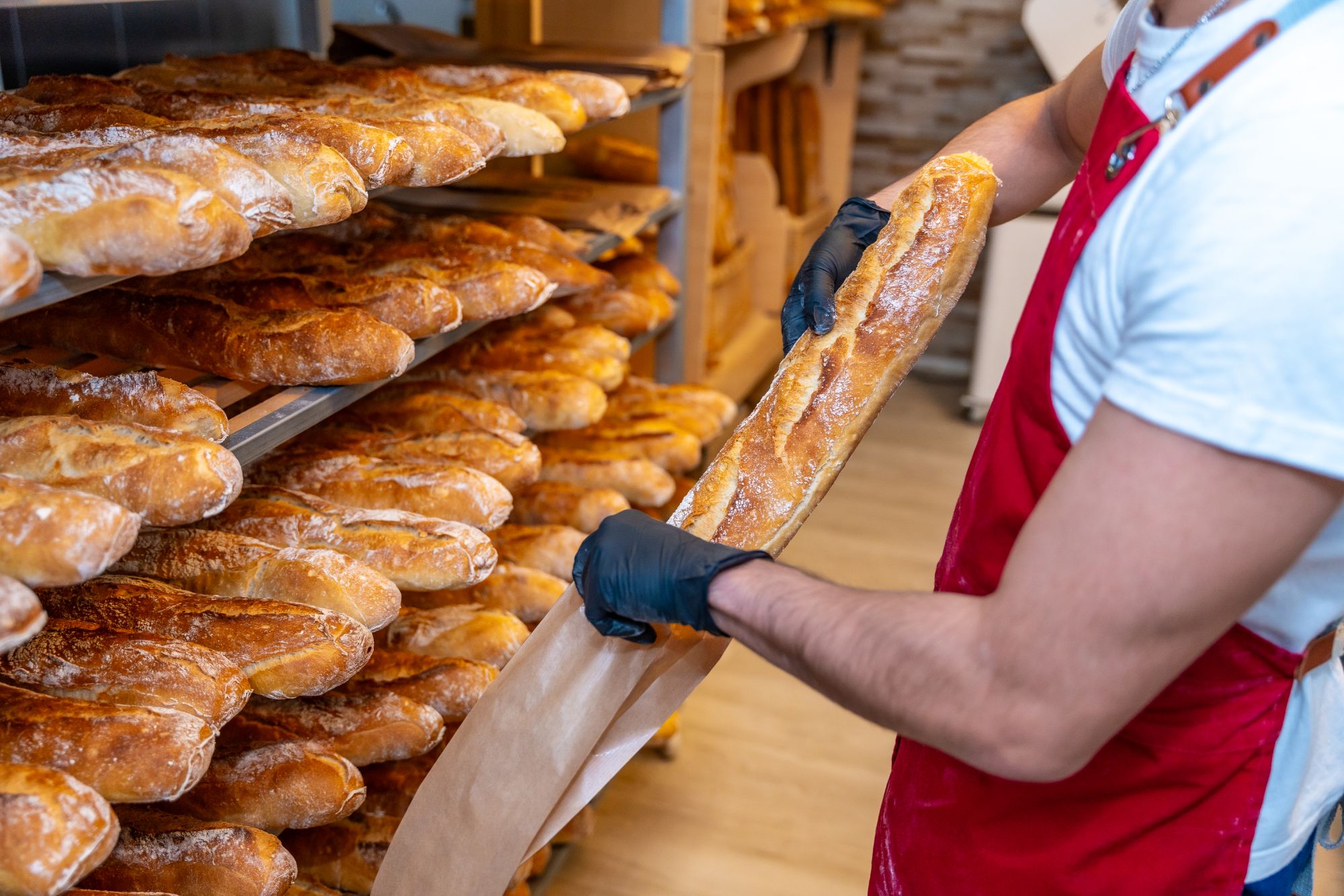 An image collage containing 1 images, Image 1 shows A bakery worker putting a baguette into a paper bag