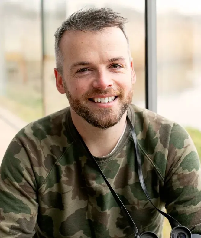 Smiling man with a beard, wearing a camouflage shirt and binoculars around his neck.