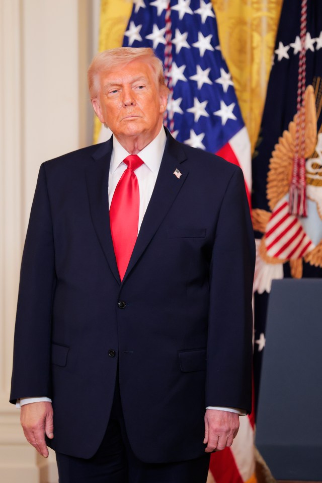 Donald Trump wearing a dark suit with a red tie and an American flag pin, standing in front of the US and presidential flags.