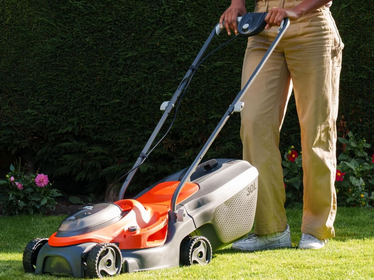 A person in tan pants and white shoes pushing a grey and orange lawnmower on a green lawn.
