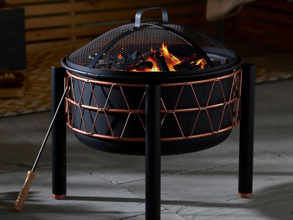 Black fire pit with copper details and a mesh lid, sitting on a patio.