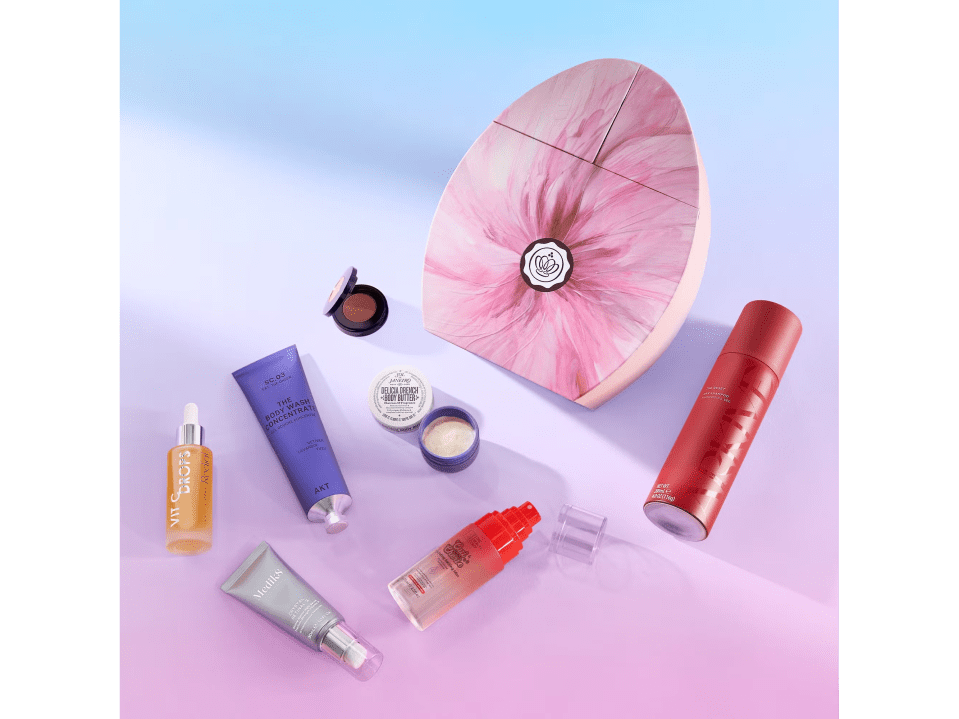 Various beauty products including a body wash concentrate, body butter, vitamin C drops, and dry shampoo, arranged around a pink floral-patterned box.