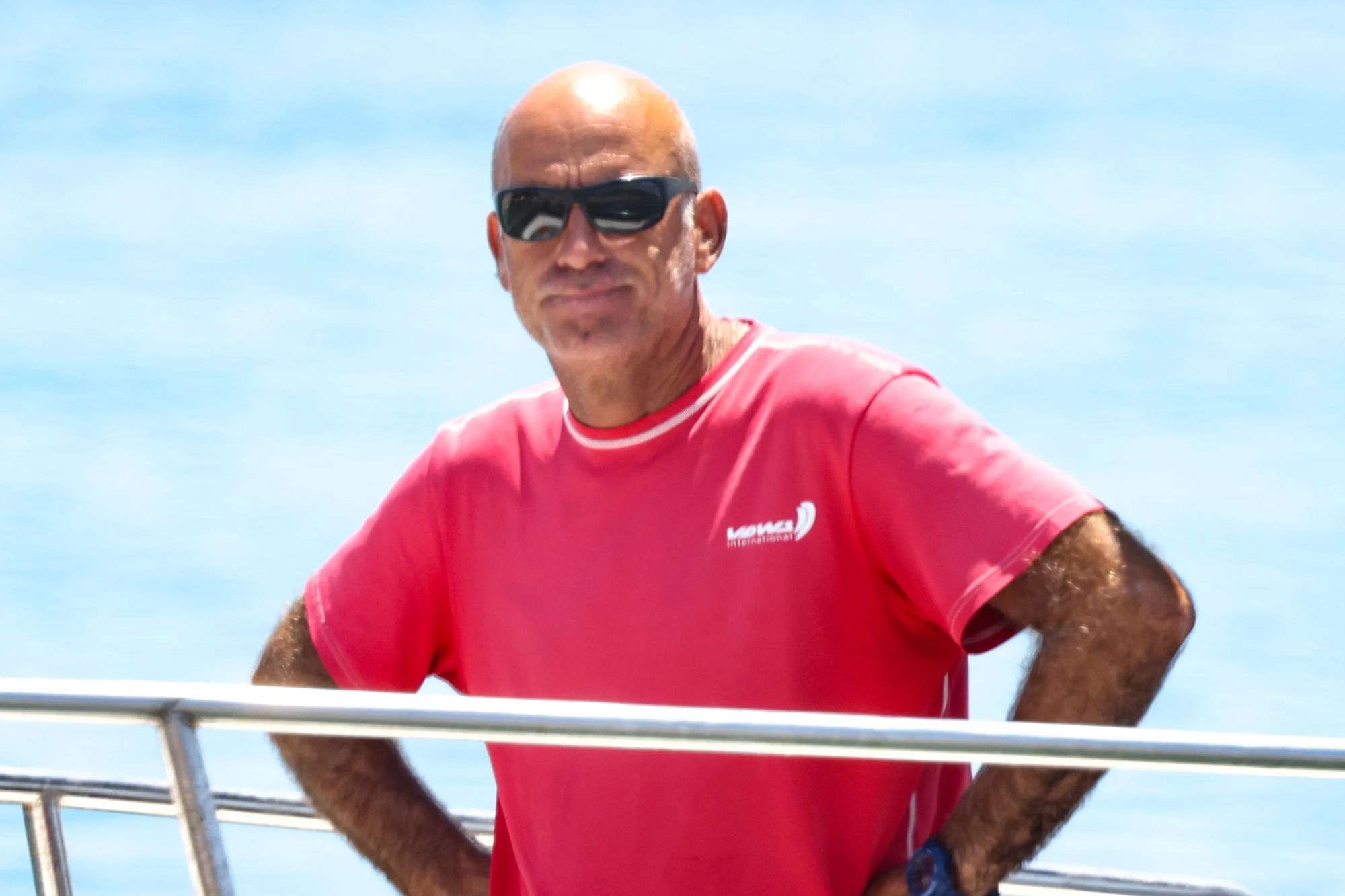 An image collage containing 1 images, Image 1 shows Nikos Mallios, wearing a red shirt and sunglasses, stands on a boat with the blue sky and sea behind him