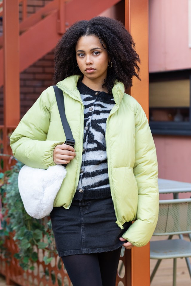 A young woman with dark curly hair and light brown skin wearing a lime green puffer jacket, a black and white striped cardigan, a black denim skirt, and black tights. She has a white fluffy bag strapped across her shoulder.