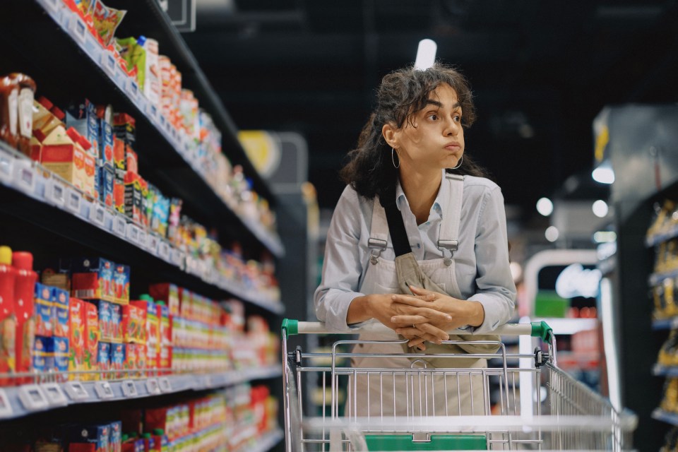 A young woman in a supermarket aisle leans on a shopping cart, looking thoughtfully at products on shelves.