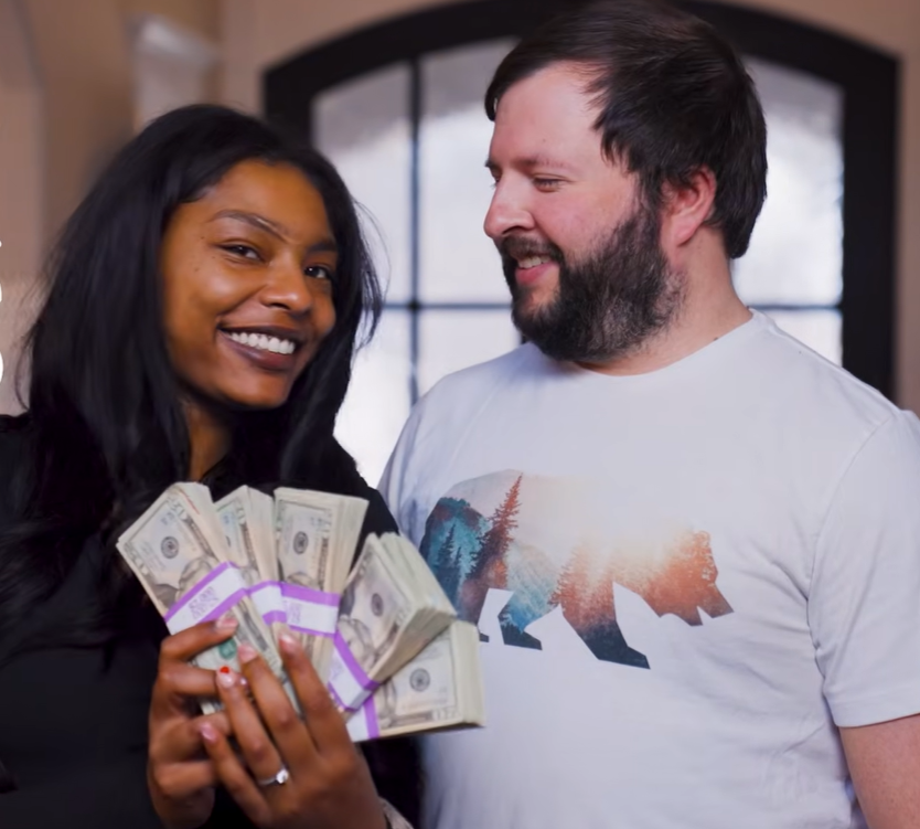 A smiling woman holds stacks of cash while a man with a beard stands beside her, looking at her.