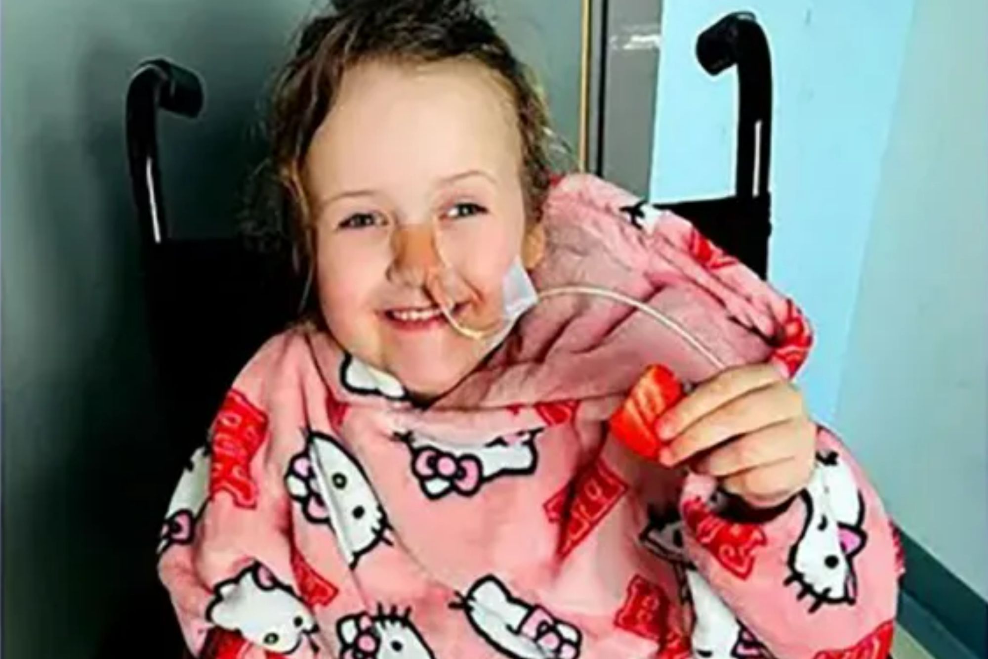 An image collage containing 1 images, Image 1 shows A young girl with a nasal cannula sits in a wheelchair wearing a Hello Kitty blanket robe and holding a strawberry