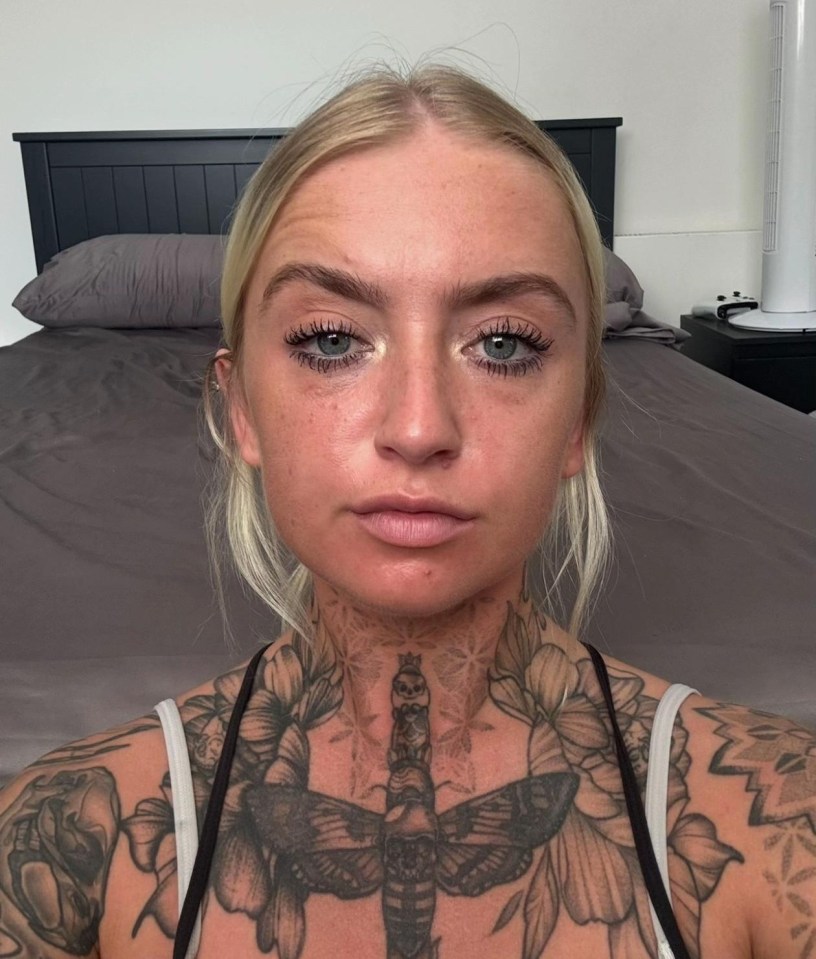 A person with heavily tattooed chest and shoulders stands before a bed.