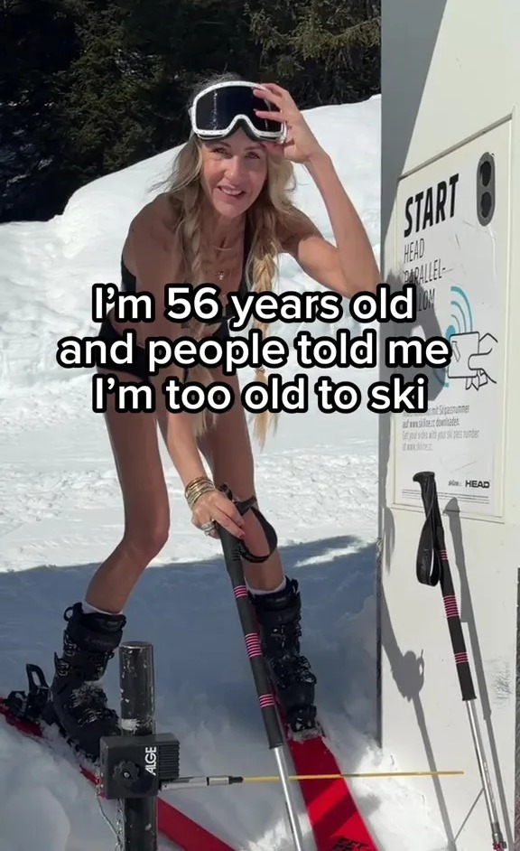 A 56-year-old woman in a bikini, ski goggles, and ski boots stands in snow with skis and poles, next to a sign that says "START," with text overlay "I'm 56 years old and people told me I'm too old to ski."