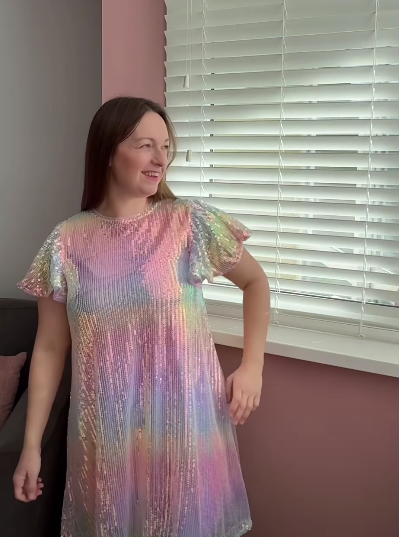 Woman in a shimmery, iridescent rainbow-colored dress smiling by a window.