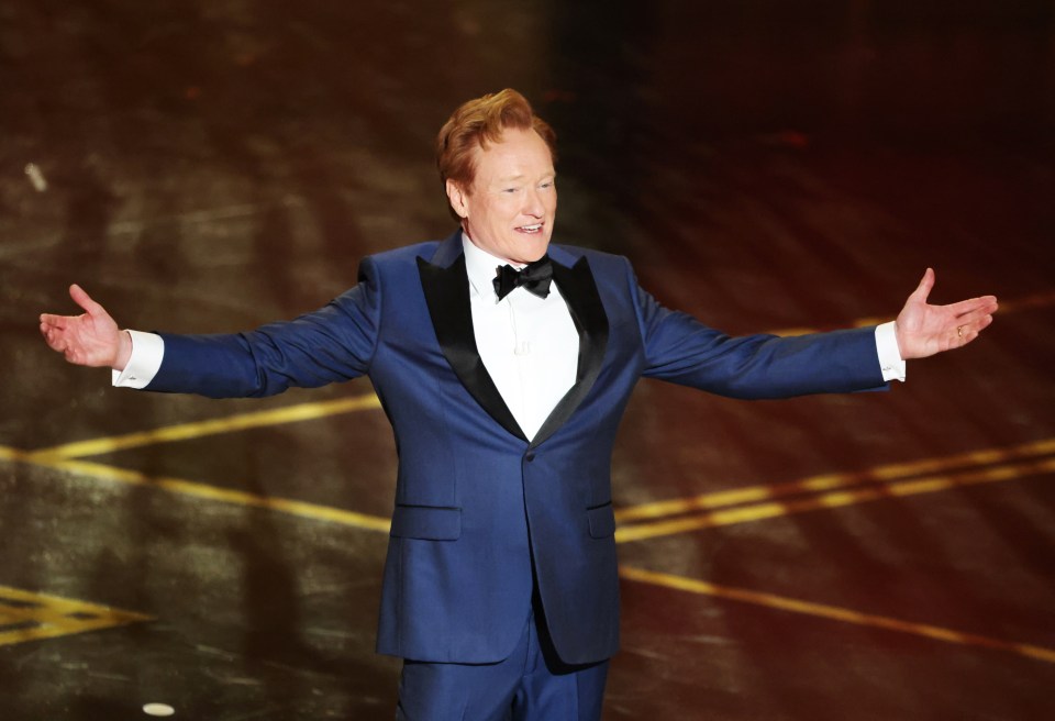 Conan O'Brien stands on stage wearing a blue tuxedo with his arms outstretched.