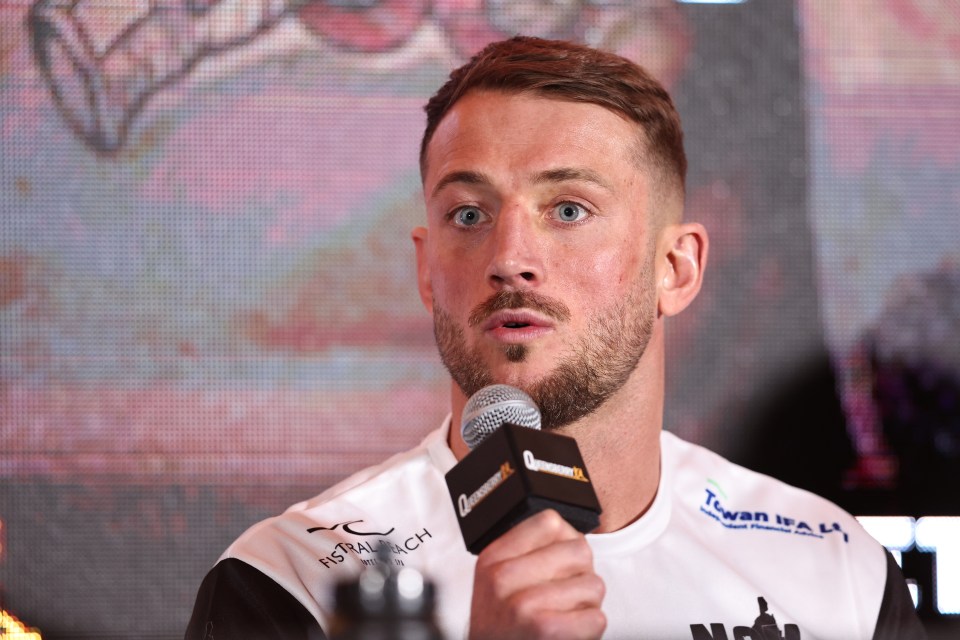 A man with light brown hair and a beard, wearing a white shirt, speaks into a microphone at a press conference.