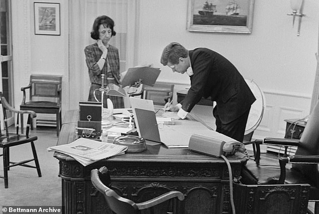 Evelyn Lincoln looks on as President Kennedy signs a document in the Oval Office